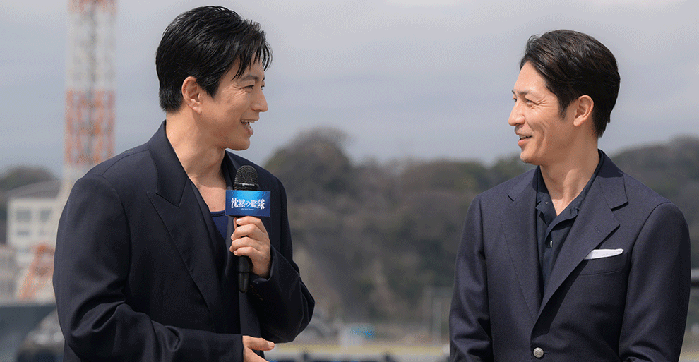 日中の屋外で、向かい合って話をする2人の男性。左側の男性がマイクを持っている。背景に木々、赤と白の電波塔のような建物。