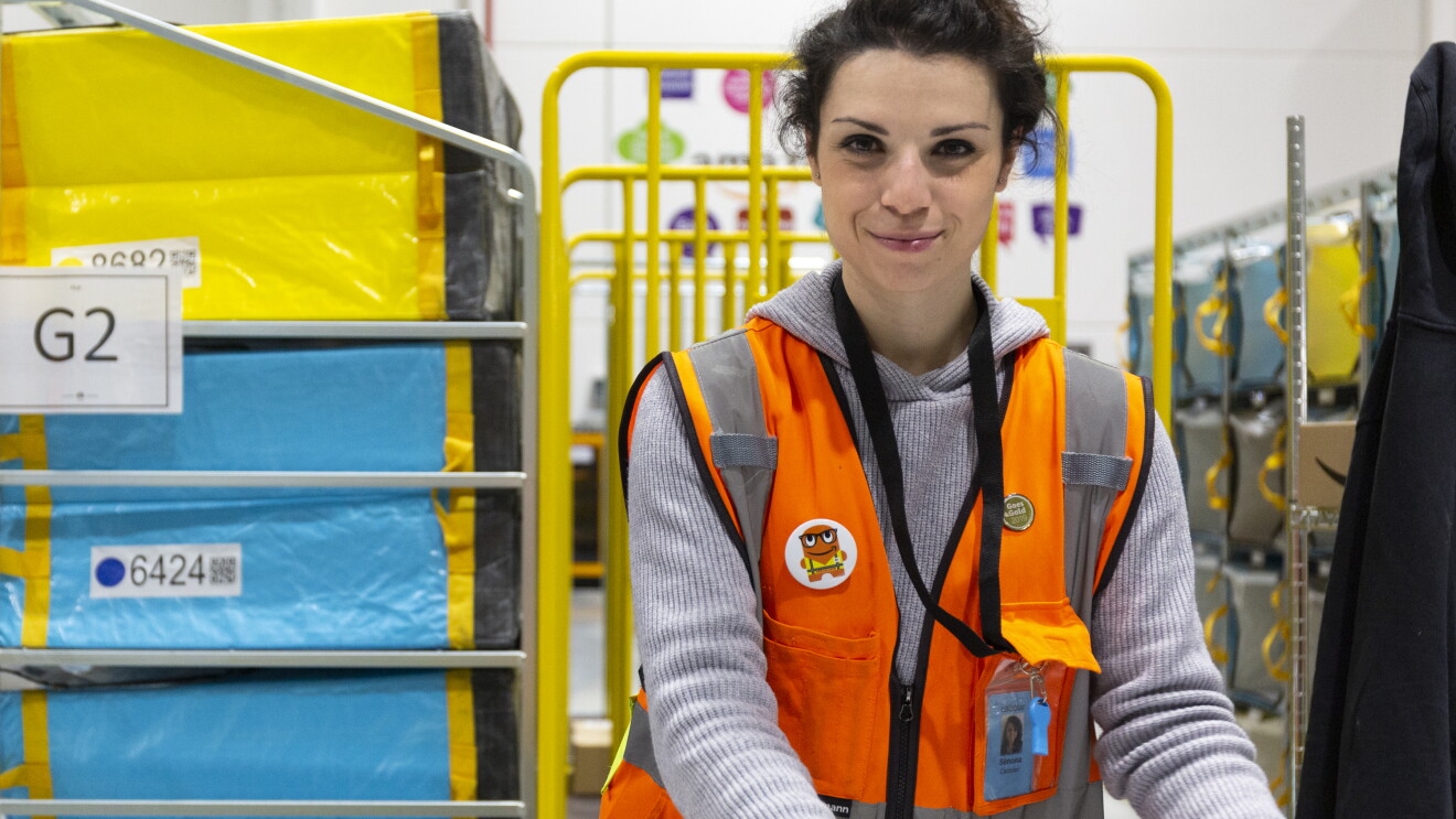 Simona Calzolari, operatrice di magazzino, inquadrata nel deposito di smistamento di Brandizzo. Indossa un giubbotto arancione ed è circondata da carrelli gialli e pacchi celesti e gialli. 