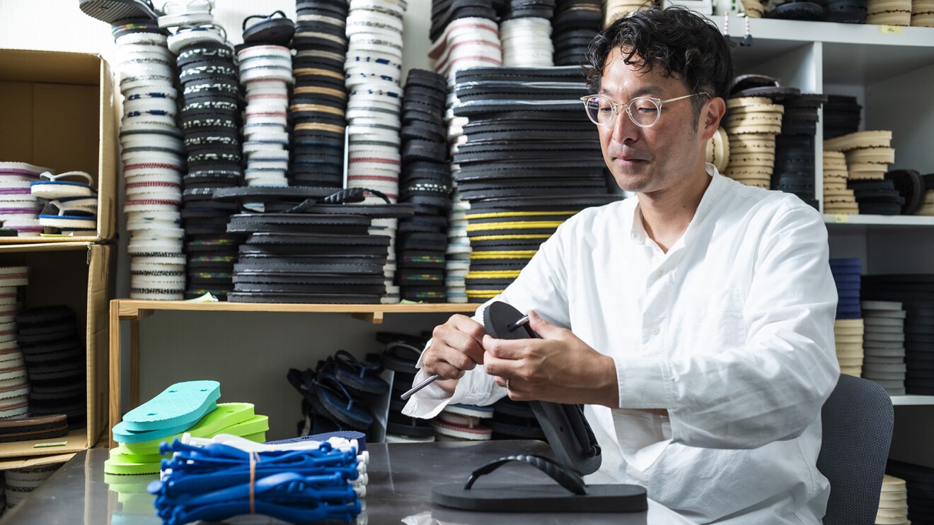 ビーチサンダルが積まれた棚の前で鼻緒をすげる作業をしているメガネをかけた男性