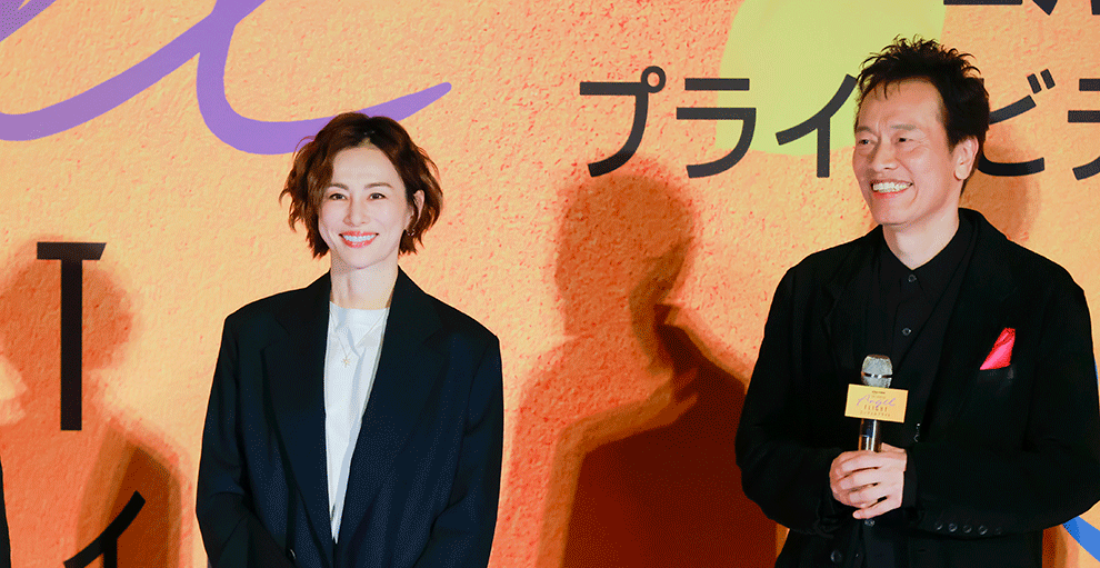 オレンジ色の壁の前、オレンジのステージの上に、黒い服を着て並ぶ2人の人物。笑顔を見せている。