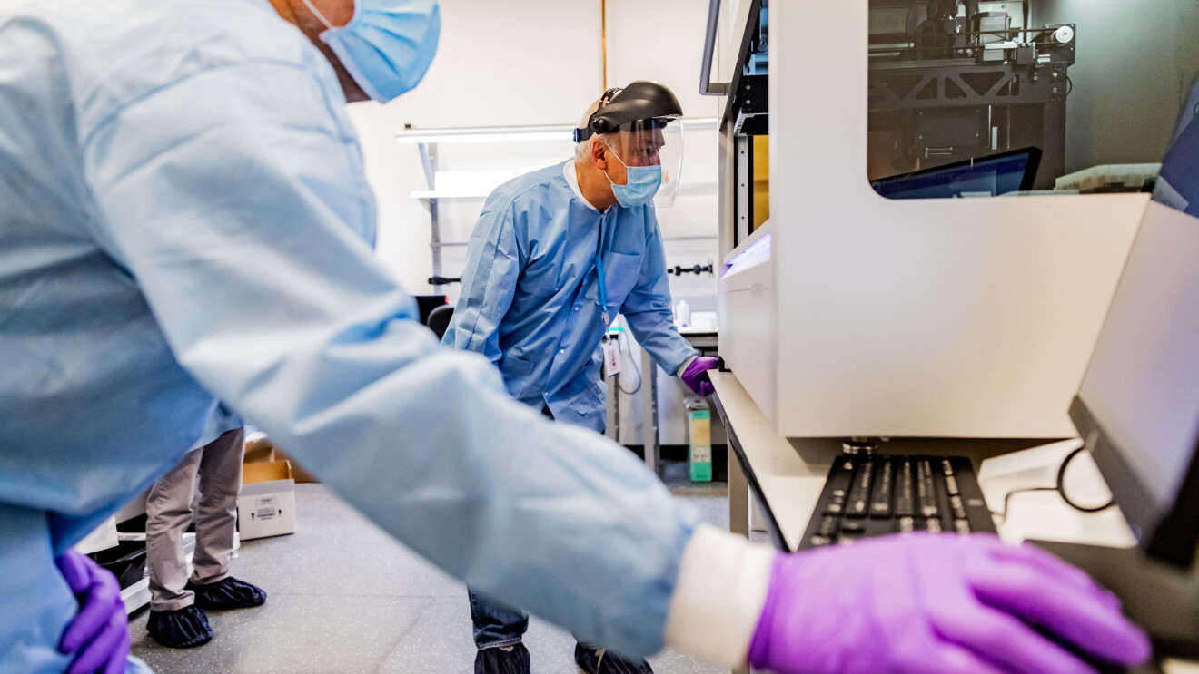 Two men working in a laboratory setting. They're wearing protective gear, including a face shield, mask, gloves, and protective clothing.