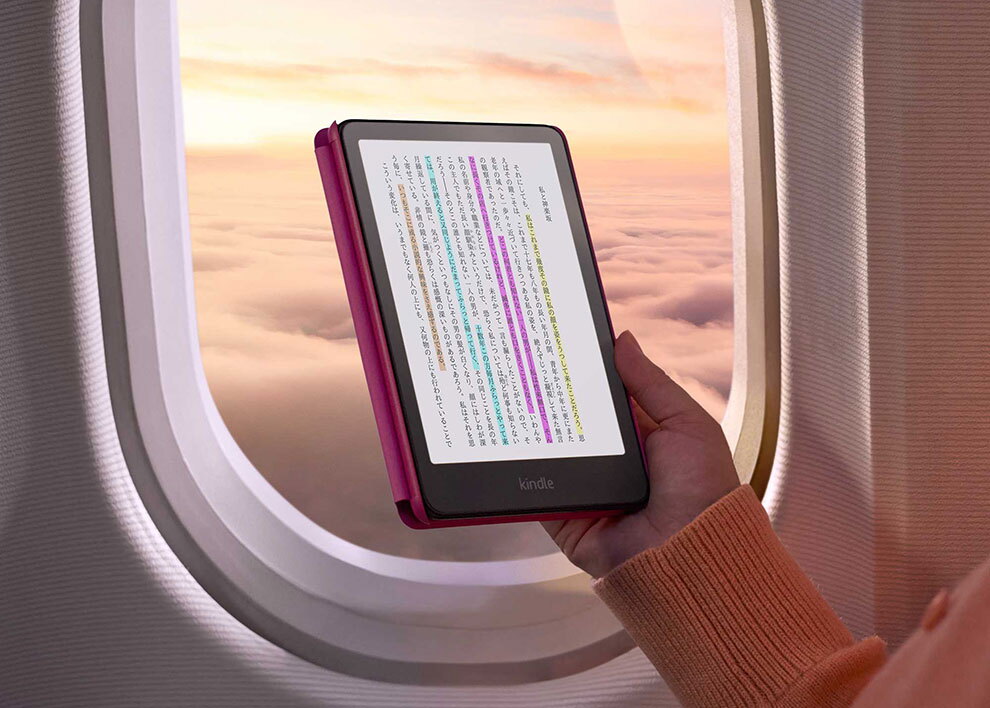 飛行機の窓際で、黒いフレームのKindle端末を片手に持っている。夕焼けの雲海が見える窓を背景に、画面には複数色のハイライトが入った縦書きの日本語ページが表示されている。