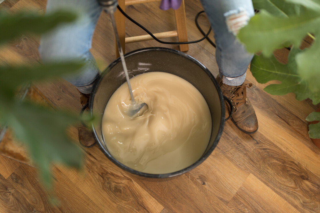 A large batch of soap is mixed inside of a large bucket, using a power drill fixed with a paddle attachment. The woman mixing the soap is wearing jeans and work boots and is seated in a chair.