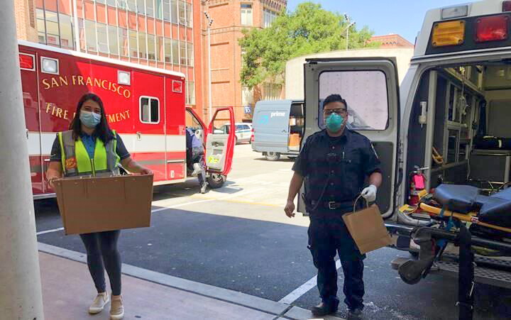 A woman wearing a safety vest and a protective mask carries a box into a fire station.
