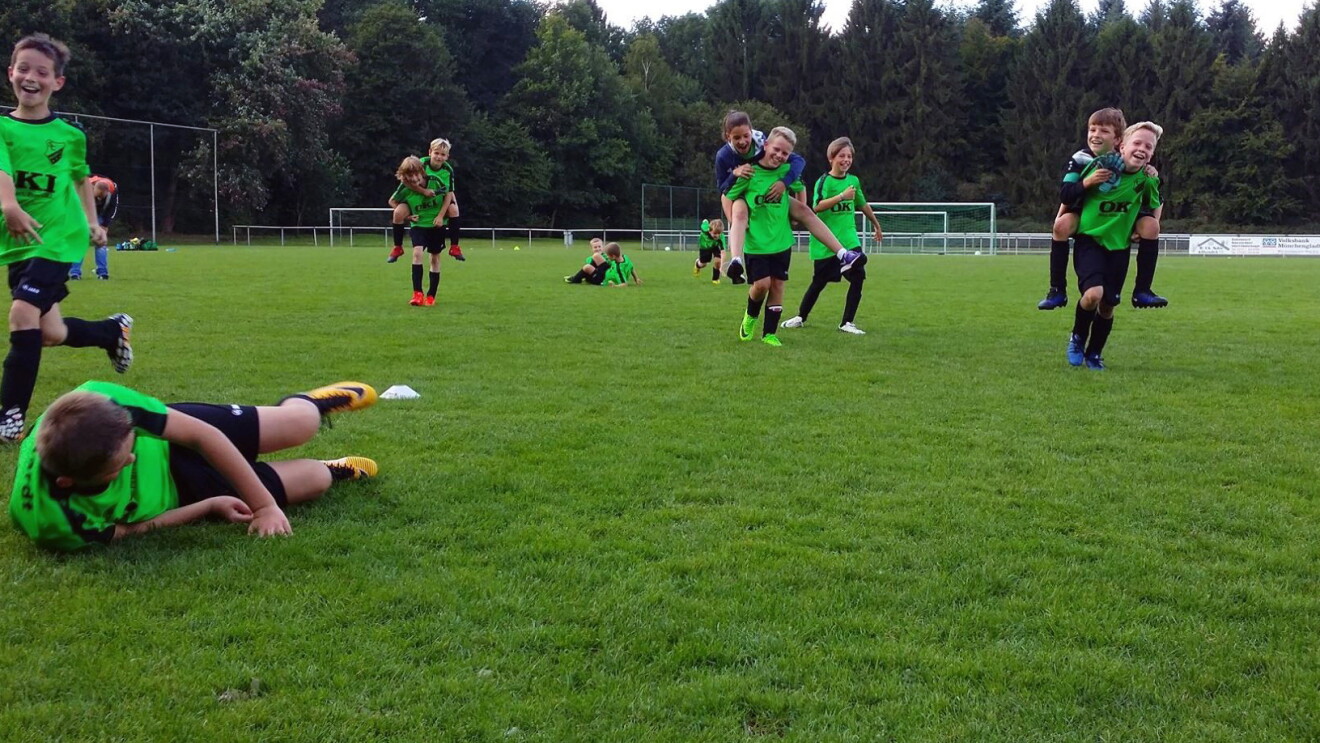 Eine Jungen-Fußballmannschaft spielt in grünen Tikots aus dem Rasen. Einige Jungen tragen ihre Freunde Huckepack, andere rennen oder sitzen auf dem Gras. Alle Kinder lachen und haben Spaß.