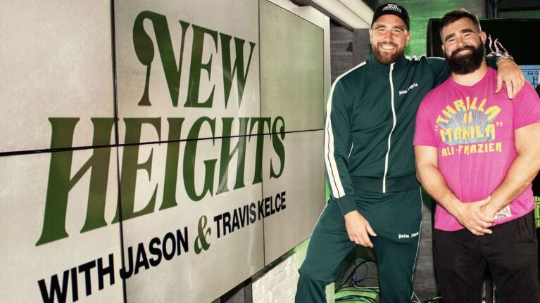 Travis and Jason Kelce standing beside 'New Heights' podcast promotional display