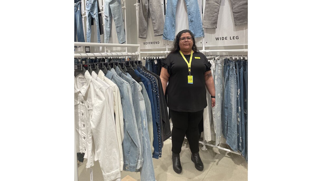 An image of a woman standing in a clothing store. Behind her is a selection of jeans on a wall in various cuts and styles.