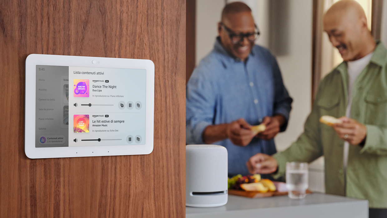 Primo piano di un device Echo Hub appeso ad un muro. Sullo sfondo, una coppia di persone sta facendo un aperitivo. 