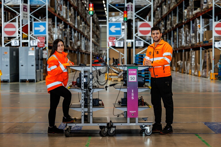 Miriam y Mario apoyados cada uno en una mesa alta transportable de Amazon. Los dos apoyan los brazo en la mesa. La mesa de él tiene un ordenador, la de ella tiene un ratón y un lector de código de barras. Los dos van con una chaqueta naranja que simula un chaleco de seguridad. De fondo el pasillo de un centro logístico. Miriam lleva el pelo largo por la cintura y es de color castaño. Él tiene el pelo corto pero la parte superior la lleva un poco más larga y la tiene rizada. Tiene los ojos claro, barba de dos días y un piercing en el labio inferior. De fondo las estanterías de un centro logístico.
