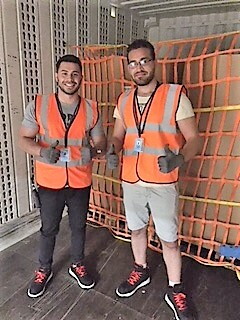 2 Versandmitarbeiter in orangenen Sicherheitswesten stehen auf der Ladefläche eines Lkws. Hinter ihnen: die mit einem Netz gesicherte Päckchen-Beladung.