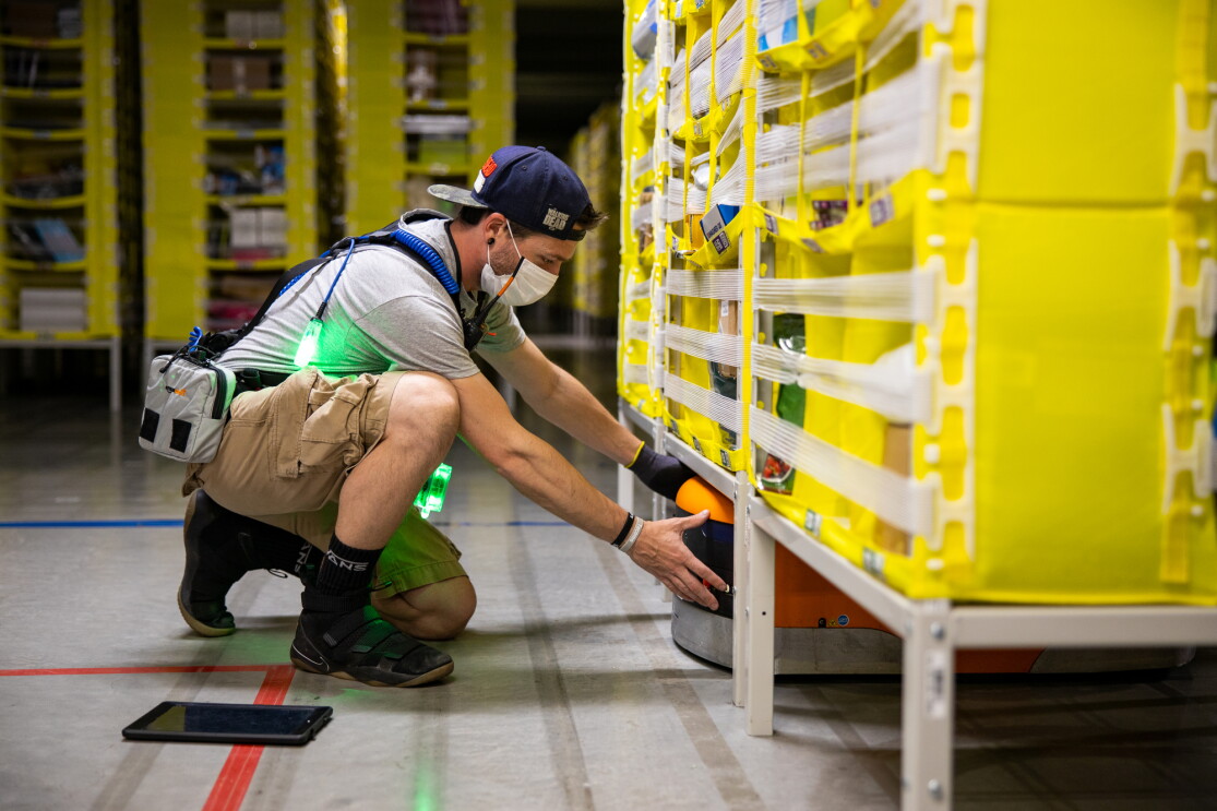 A man at an Amazon fulfillment center neals in front of a yellow storage tower, reaching under to maneuver an orange robot.
