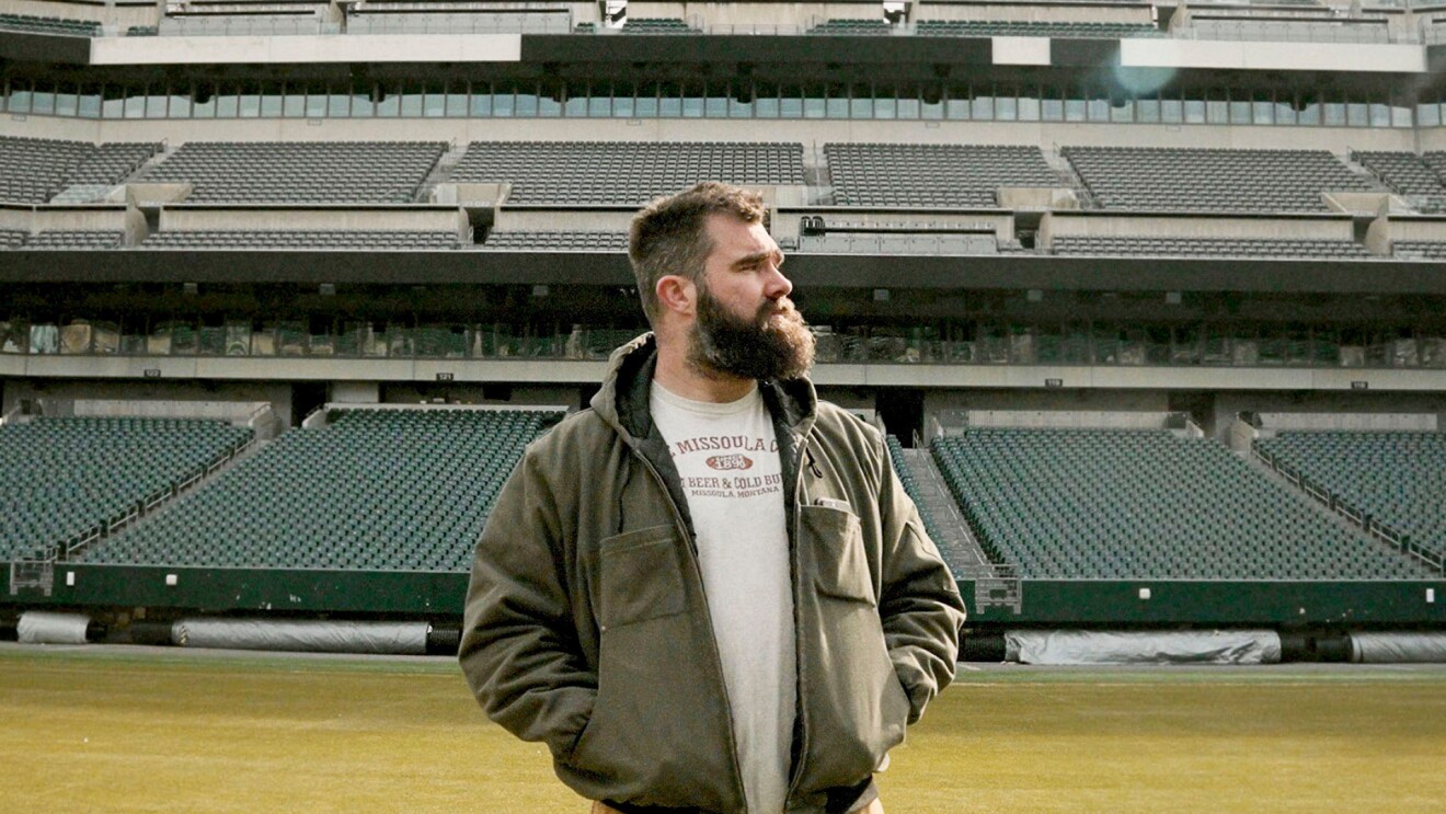 Kelce stands in the middle of a football field in an empty stadium.