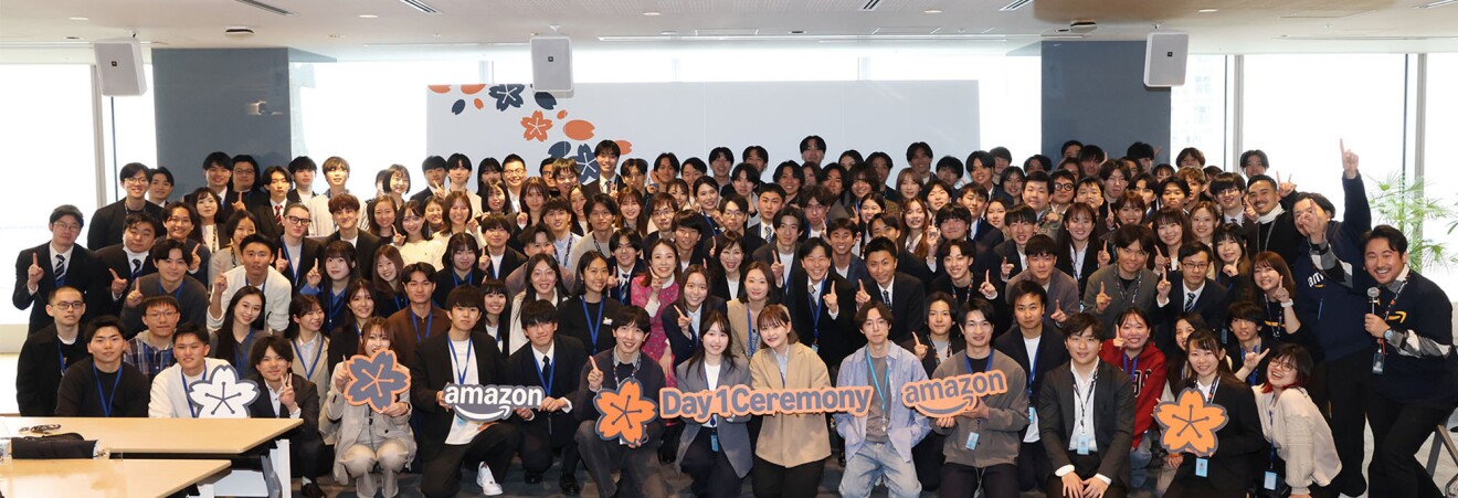 明るい会議室に集まっている100人以上の人々一番前の人たちはDay 1 ceremonyと書かれたオレンジ色のパネルを持っている