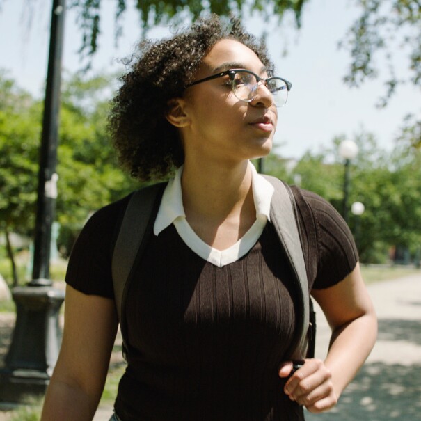 nicole, an amazon future engineer scholarship recipient, walks down a campus road while wearing a backpack. she's wearing glasses and is looking off camera to her left