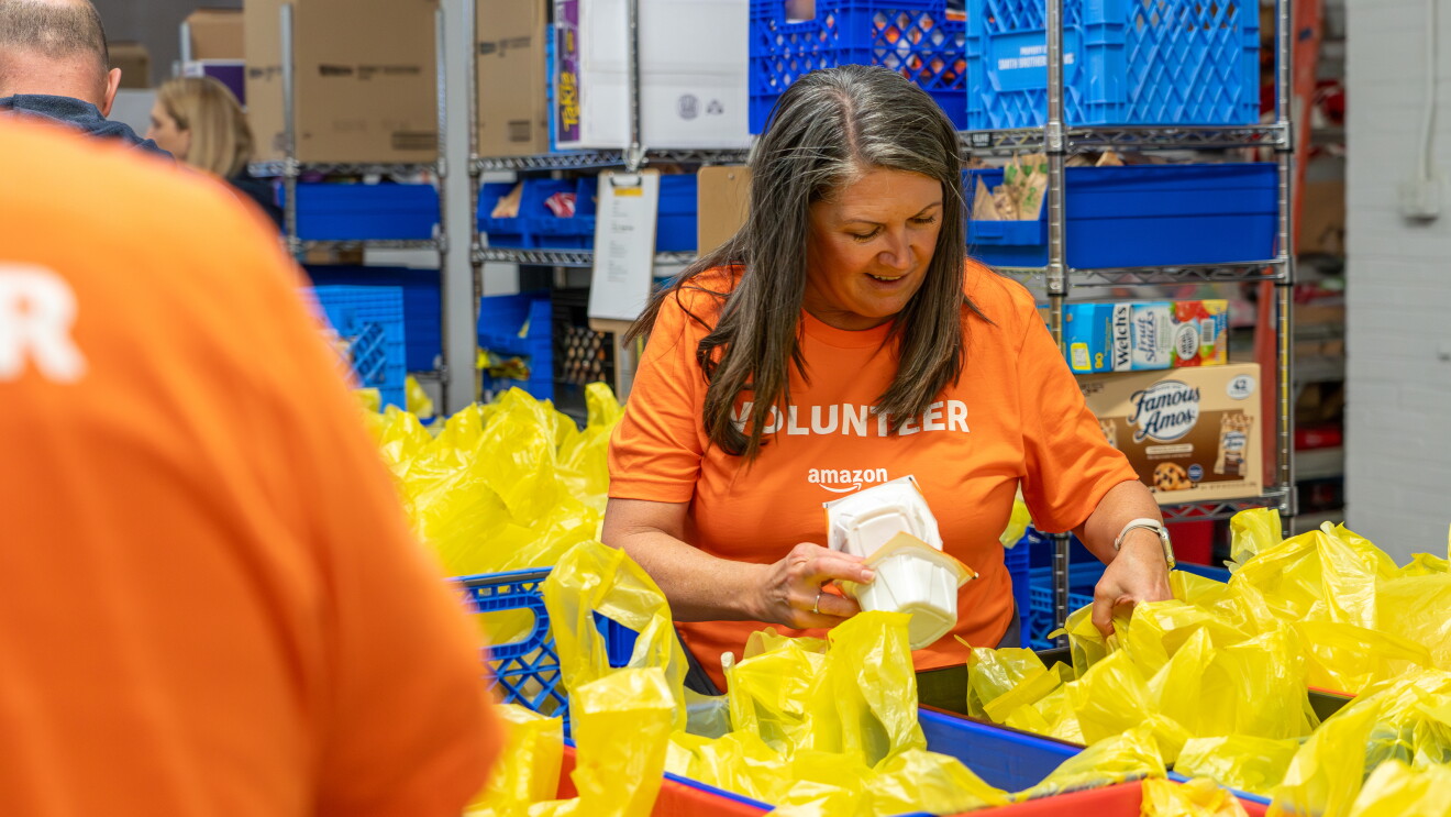 Amazon volunteer sorting food items into bright yellow bags at a food bank warehouse
