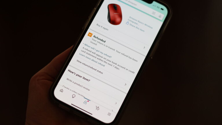 An image of a hand holding a phone. On the screen is text confirming the return of an item on Amazon and an image of the product. It's a red computer mouse.