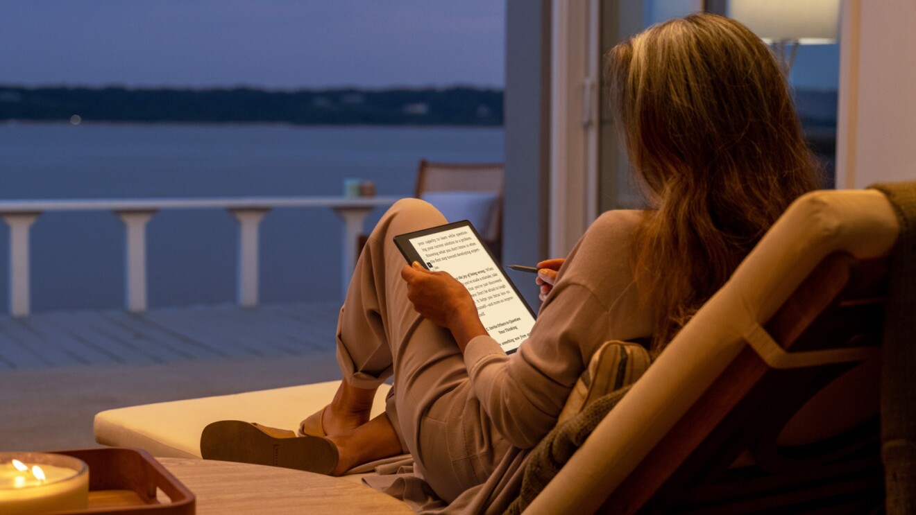 An image of a woman sitting on a patio chair by the water with a lit candle next to her reading from a Kindle Scribe. 