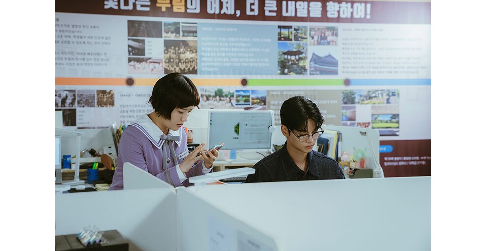 パーテーションで区切られたデスクが配置された室内、机に向かう男性とその横側に立ってスマホを操作する少女。背景にハングル文字と画像。