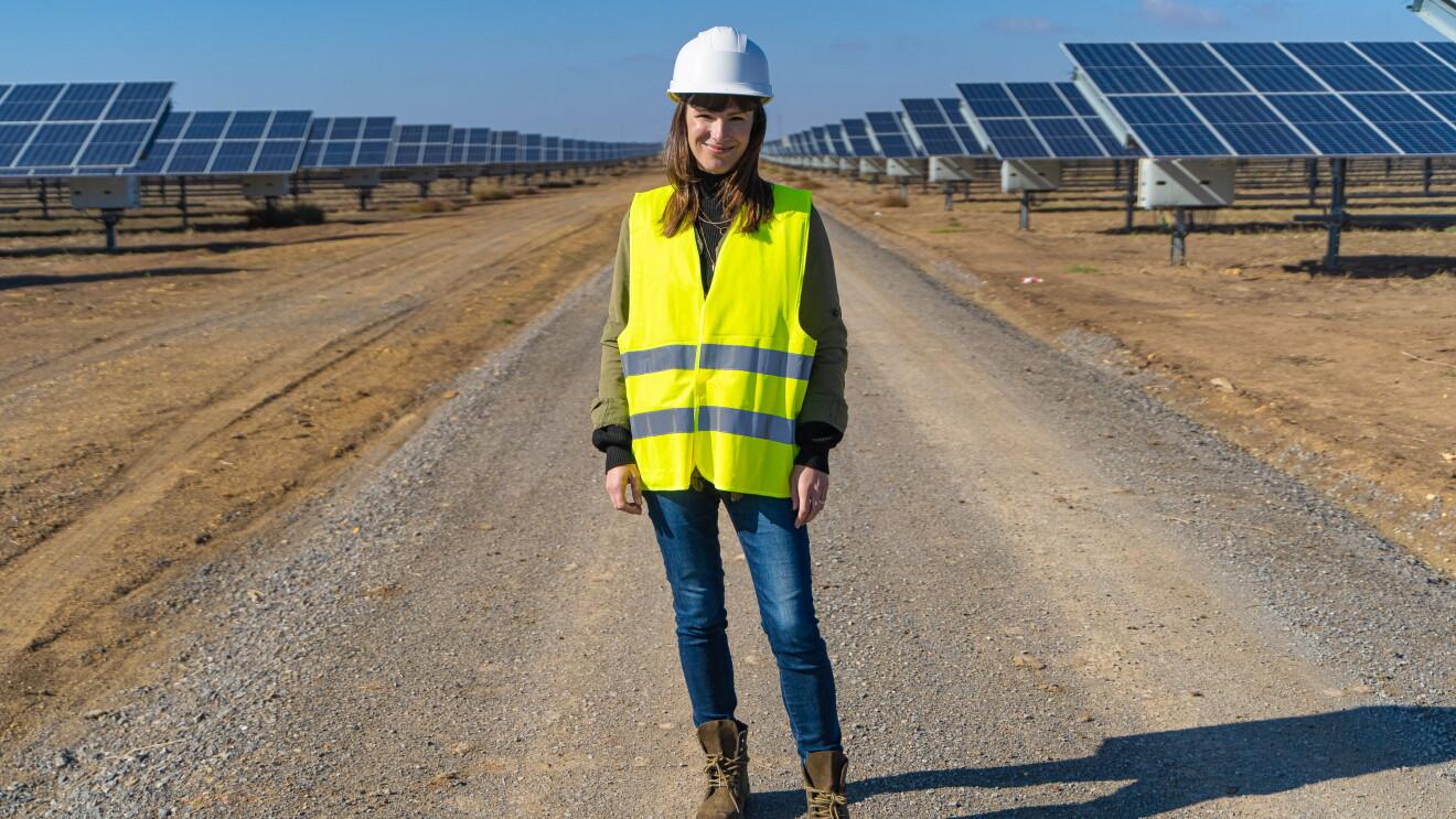 Denise Hugo, jefa de proyectos de Energías Renovables de Amazon. Ella está en medio de la foto, en un camino de tierra y entre dos líneas de placas solartes. Va vestida con unas botas marrones, unos vaqueros, casco blanco y chaleco amarillo.
