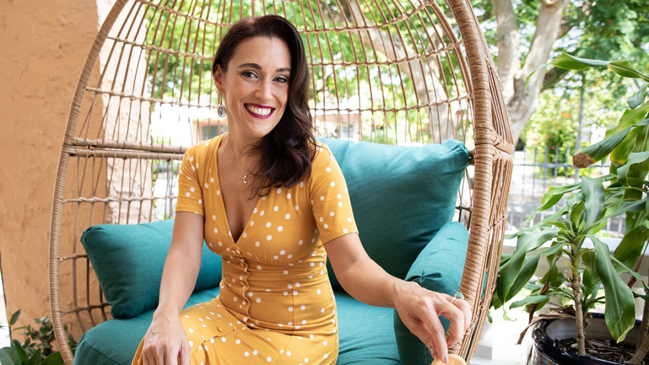 An image of a woman sitting in a wicker chair outside while smiling for a photo in a yello dress with white polka dots. The decor around her is colorful.