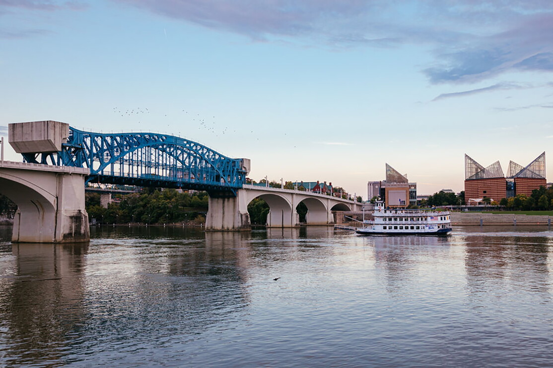 A sweeping view of a river, with a bridge stretched across the width of the body of water. A riverboat is seen in the background, in front of architecturally interesting buildings.