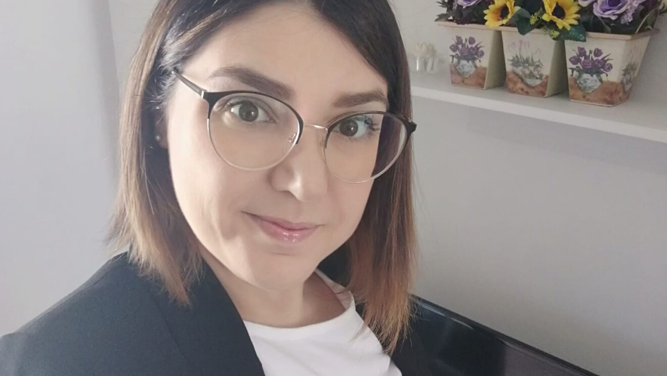 Primo piano della volontaria Amazon Francesca. La donna ha i capelli a caschetto, indossa degli occhiali da vista tondi, una giacca nera e una maglia bianca. Alle sue spalle degli scaffali su cui sono poggiati vasi con girasoli.