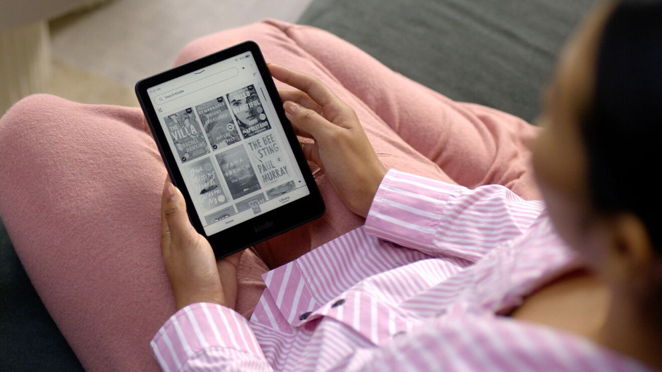 Person reading on an Amazon Kindle displaying book covers while relaxing on a pink cushion