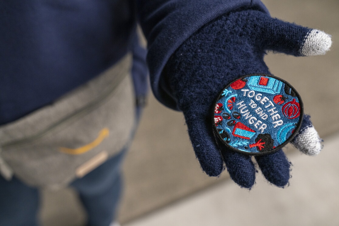 A closeup of a gloved hand holding the Together to End Hunger badge, designed by artist Stevie Shao.