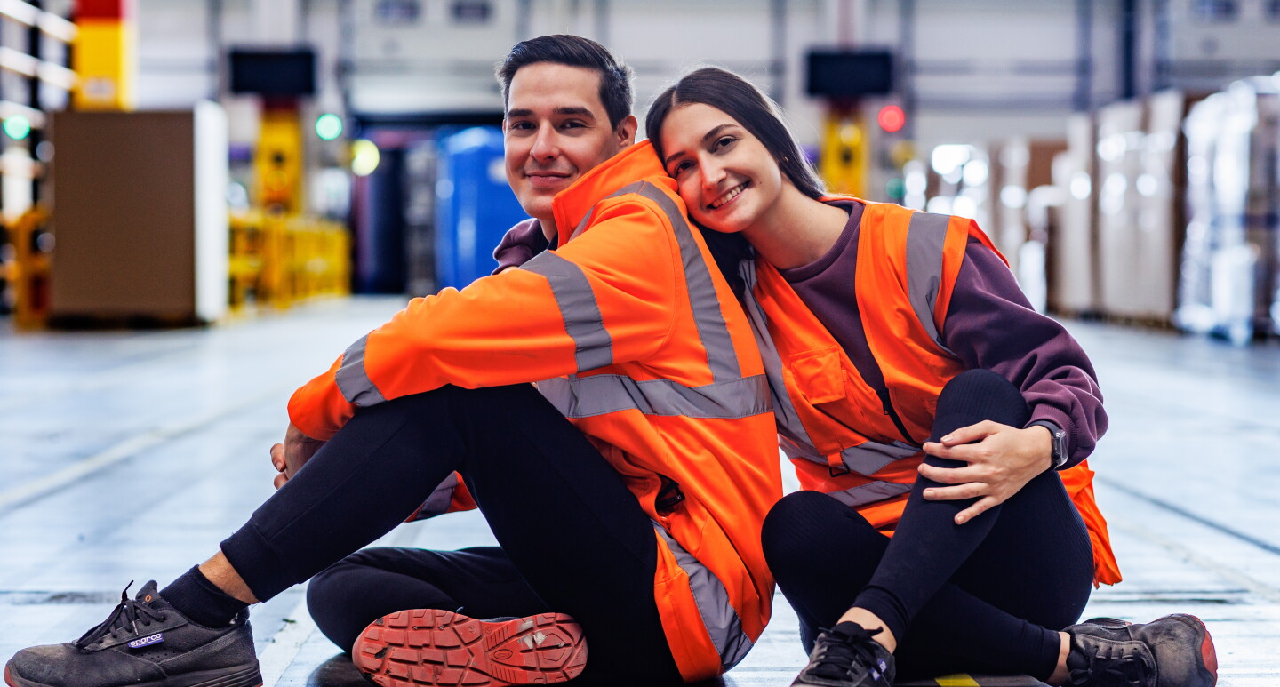 Fabiana y Rafael posando en el suelo de un centro logístico de Amazon. Los dos están posando, mirando de frente a cámara. Ella está de frente, los las piernas cruzadas, zapatos de seguridad negros, pantalón negro, camiseta morada y chaleco naranja. Lleva el suelo y es lacio y largo, Agarra con la mano derecha a Rafael. Él está de lado con las piernas cruzadas pero mirando a cámara. Lleva también zapatos de seguridad, pantalón negro, chaqueta que imita al chaleco de color naranja.