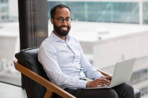 An image of a man seated in front of an office building window with a laptop.