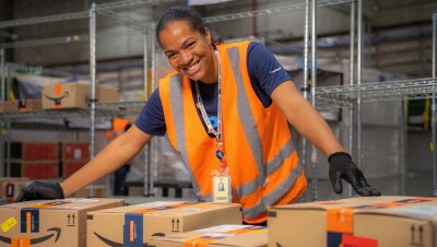 Uma mulher mexe em caixas de entrega da Amazon em um centro de distribuição. Ela usa luvas pretas e um colete laranja