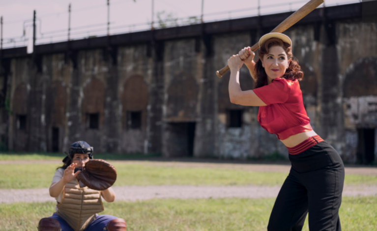 D'Arcy Carden holds a bat on a baseball field in A League of Their Own