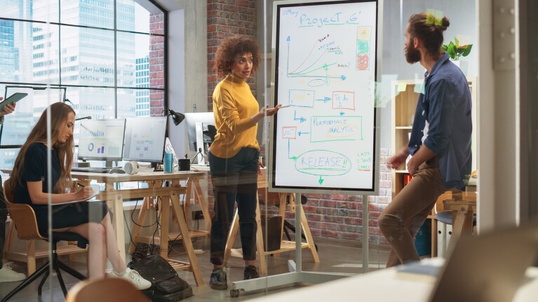 A photo of a man and woman standing in front of a white board discussing content written on the board. They are in a conference room where there are two additional people writing notes.