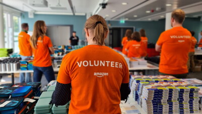 Man sieht Amazon Mitarbeiter:innen mit orangenem Volunteer-T-Shirt von hinten. Sie stehen hinter Tischen mit Schulmaterialien.