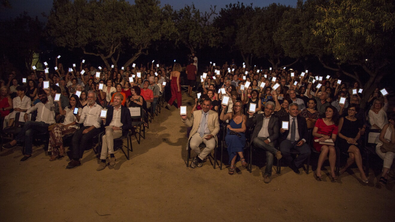 Platea di persone sedute nella Valle dei Templi di Agrigento durante un reading di Luigi Pirandello in occasione dei 150 della sua nascita. Gli spettatori reggono con una mano un Kindle PaperWhite, con lo schermo rivolto verso la camera. Sullo sfondo, degli alberi. 