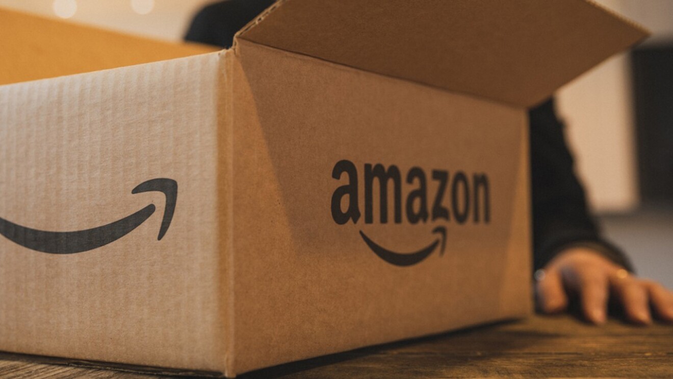 Primer plano de una caja de Amazon abierta encima de una mesa. Se aprecian dos de las cuatro caras de la caja. En una se ve la sonrisa y en la otra Amazon y la sonrisa. La caja es de cartón y está abierta. La mesa es de madera de color marrón. Detrás de la caja hay una mano apoyada a la mesa. Se ven cuatro dedos y un anillo. La persona lleva un jersei negro de manga larga. Se le ve el brazo izquierdo y un trozo de hombro derecho.
