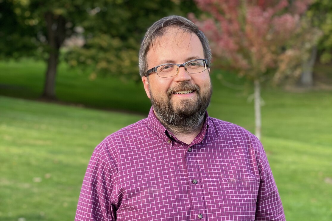 A man smiles at the camera. He wears glasses and a button down, and stands in a park setting.