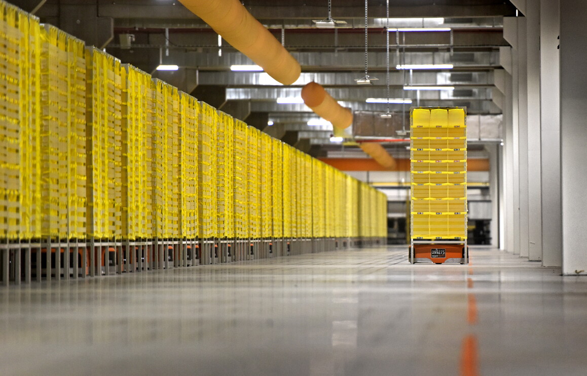 Ein Roboter transportiert ein gelbes Regal durch eine Halle. Links im Bild sind in einer langen Reihe weitere gelbe Regale zu sehen unter den Transportroboter sind.
