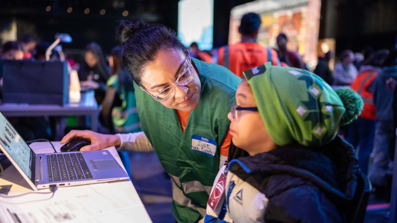 Amazon’s Winter Wonderland: Volunteer in green vest assists young participant with laptop at tech event
