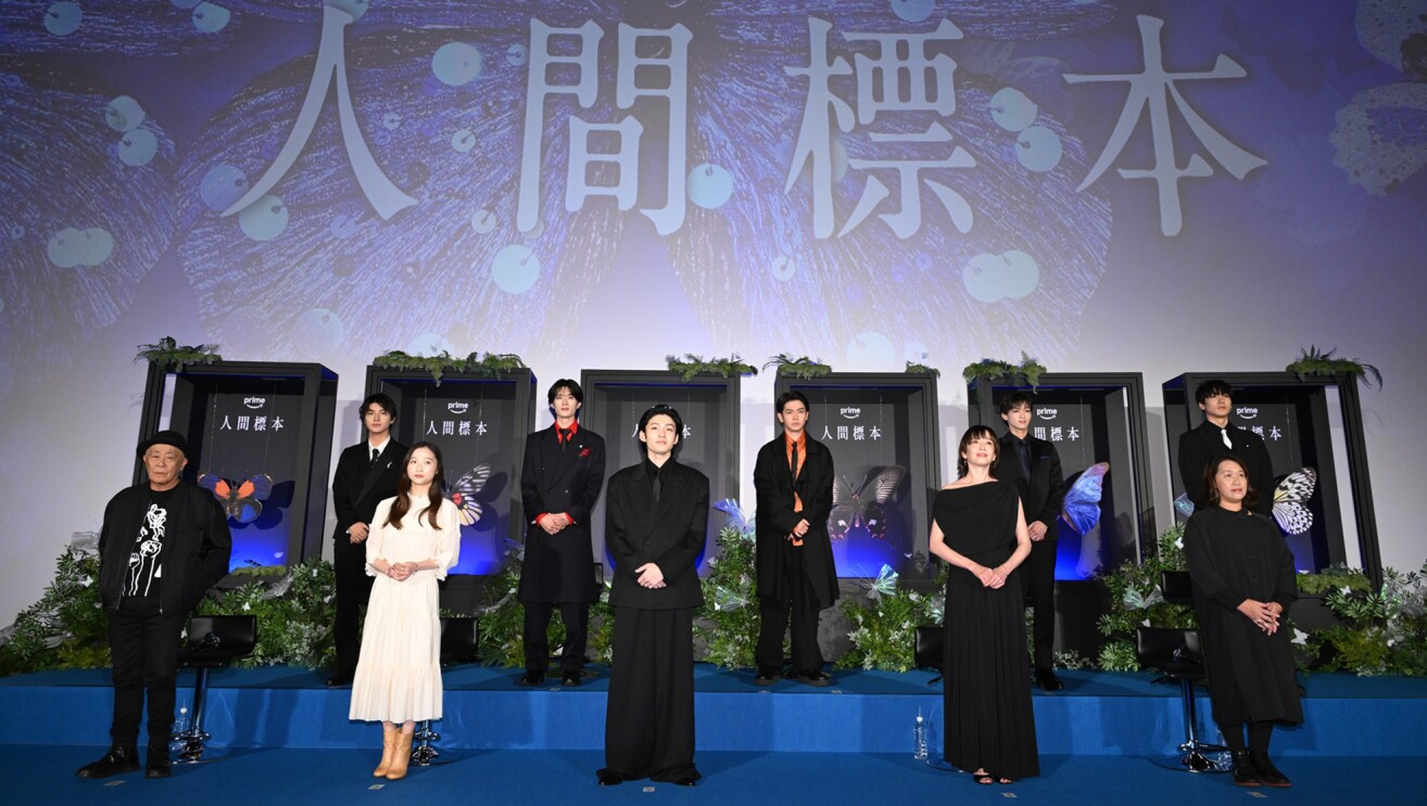 青いステージに2列に並んで立つ10人の人物。前列左から2番目の女性以外、全員黒ずくめ。ステージ上の壁に「人間標本」の大きな文字。