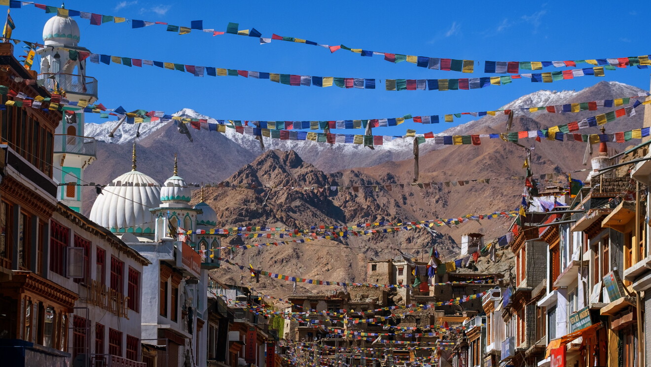 Under a bright blue sky and colorful prayer flags, multi-story buildings with shops are photographed on facing sides of a street. Domed structures and snow-capped mountains are in the background.