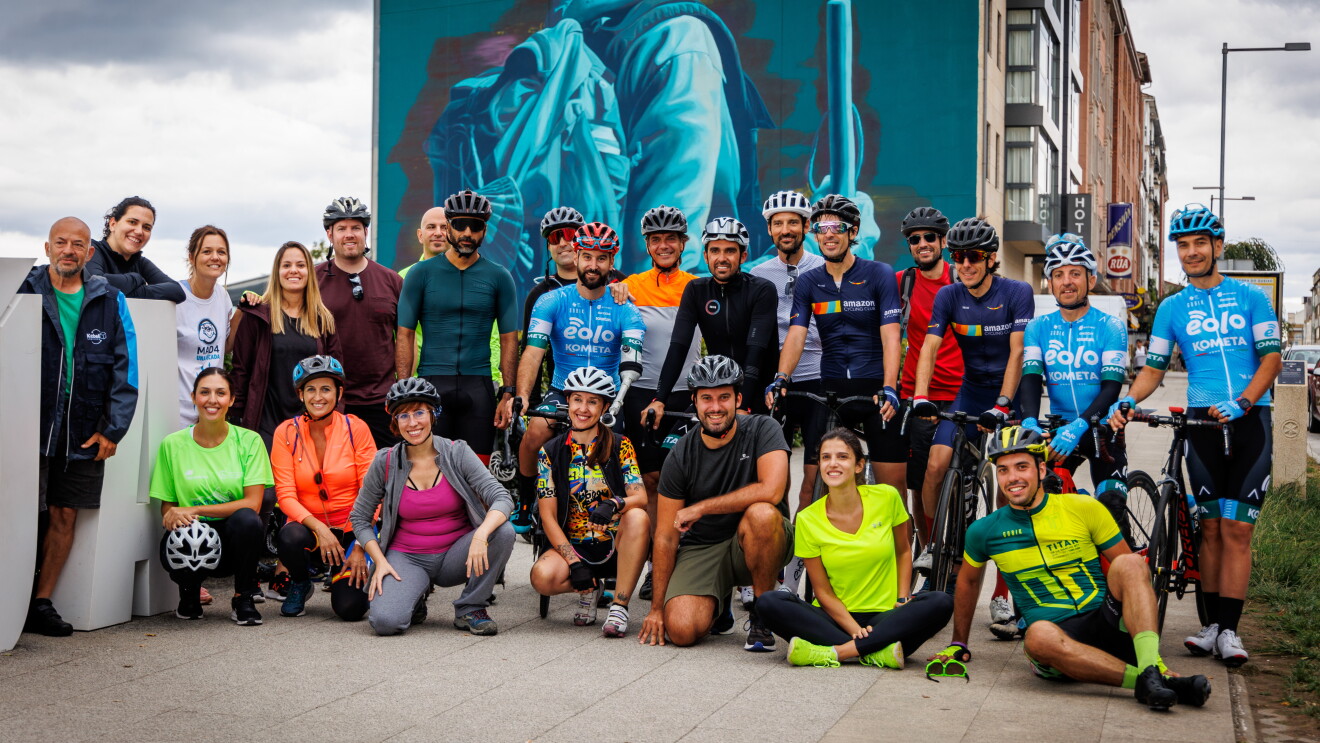Una foto de grupo con 24 personas entre hombres y mujeres posando para una foto de equipo. Algunos están sentado en primera fila y los de segunda fila algunos están apoyados en la bicicleta. Algunos llevan el casco puesto y otros no. De fondo se ve la pread de una edificio que tiene un graffiti pintado de tonos azules con una persona caminando y el logo y las letras de Xacobeo 2021. 