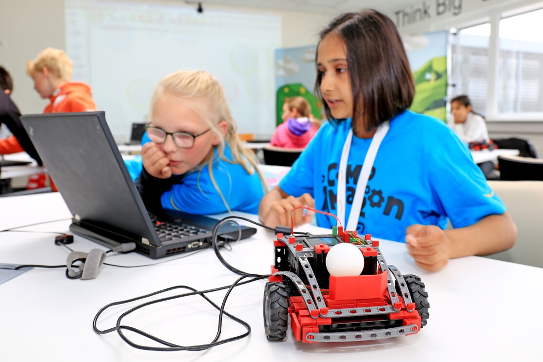 Ein rotes kleines Fischertechnikfahrzeug steht auf einem Tisch. Es enthält einen weißen Tennisball. Im Hintergrund sind zwei Mädchen im blauen Shirts zu sehen, sie blicken intensiv in einen Laptop
