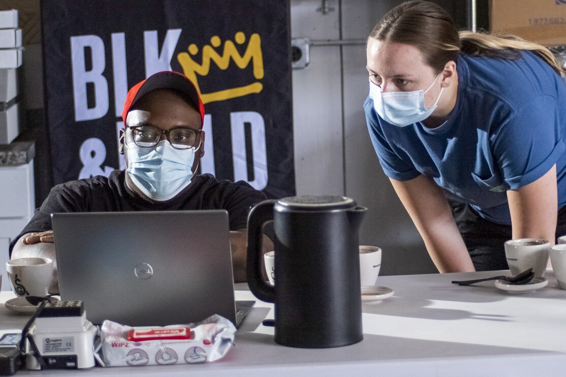 One person works on a laptop while another leans in and looks on.