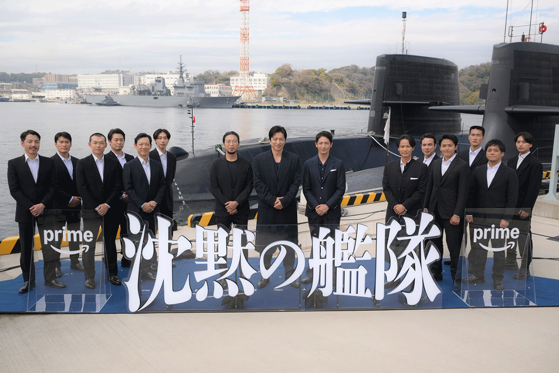 接岸された潜水艦を背に、港に並ぶジャケット姿の男性たち。中央に3人、左右に6人ずつ立っている。背景の水面には船が2隻浮いている。
