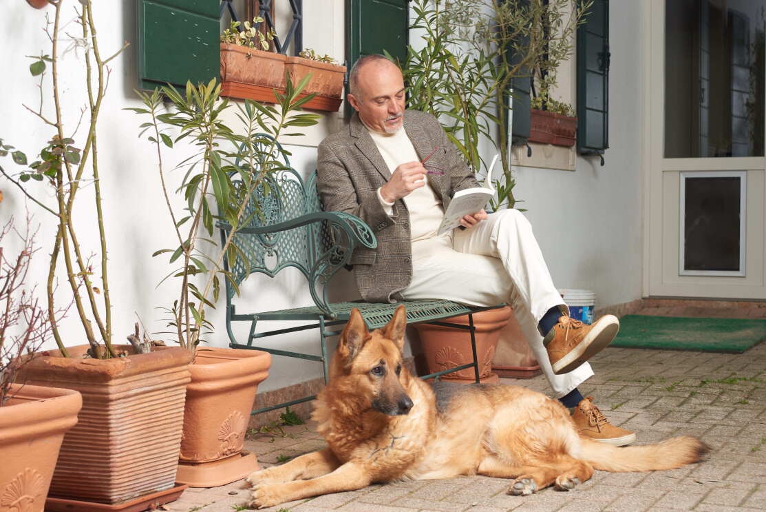 Lo scrittore Federico Maria Rivalta legge un libro sul portico di casa con un pastore tedesco ai suoi piedi.