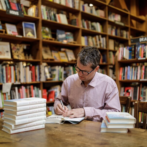 An author sits at a table and signs his book in a bookstore, surrounded by shelves of other book titles.