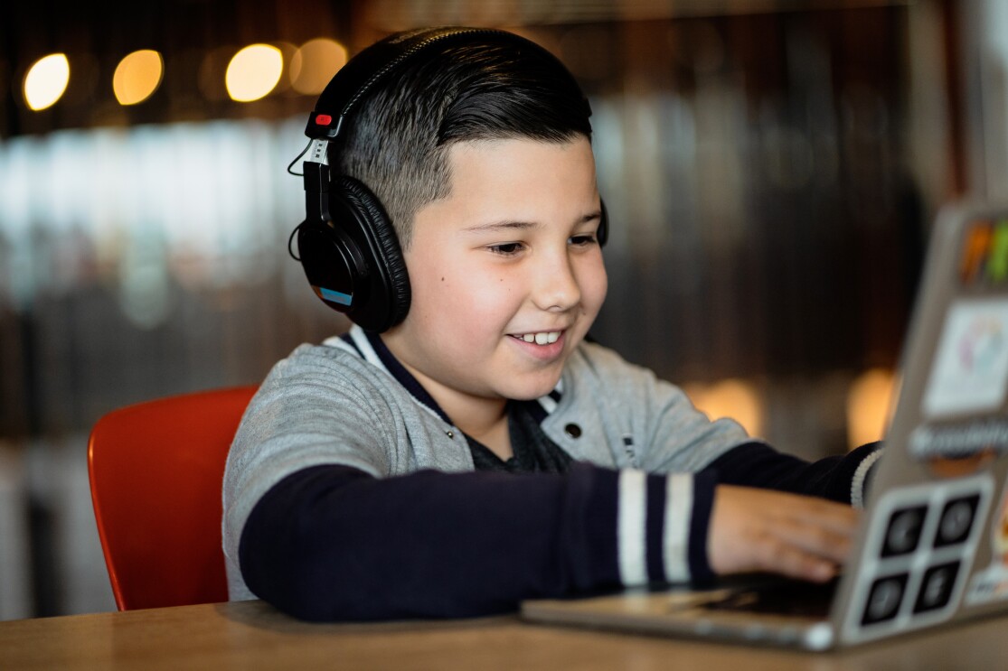 A boy wearing headphones works on a laptop at a coding camp while smiling.