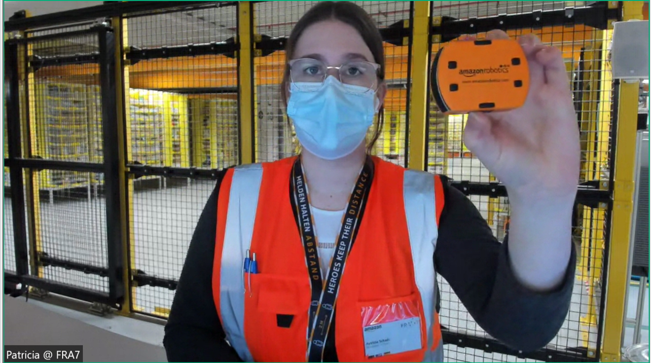 Patricia Schade, Tourleiterin bei Amazon und Verantwortliche des Besucherprogramms Deutschland stet in einem Logistikzentrum und hält iein oranges Teil in die Kamera, auf dem Amazon Robotics steht. Sie trägt eine orange Warnweste, ein Headset und einen Mundschutz.