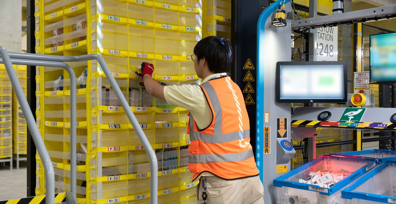 物流センターの作業ステーション。オレンジのベストと赤い手袋を着用したスタッフが、細かく区切られた黄色い商品棚から商品をピッキングしている。右側には複数のモニターと半透明の収納ボックス。